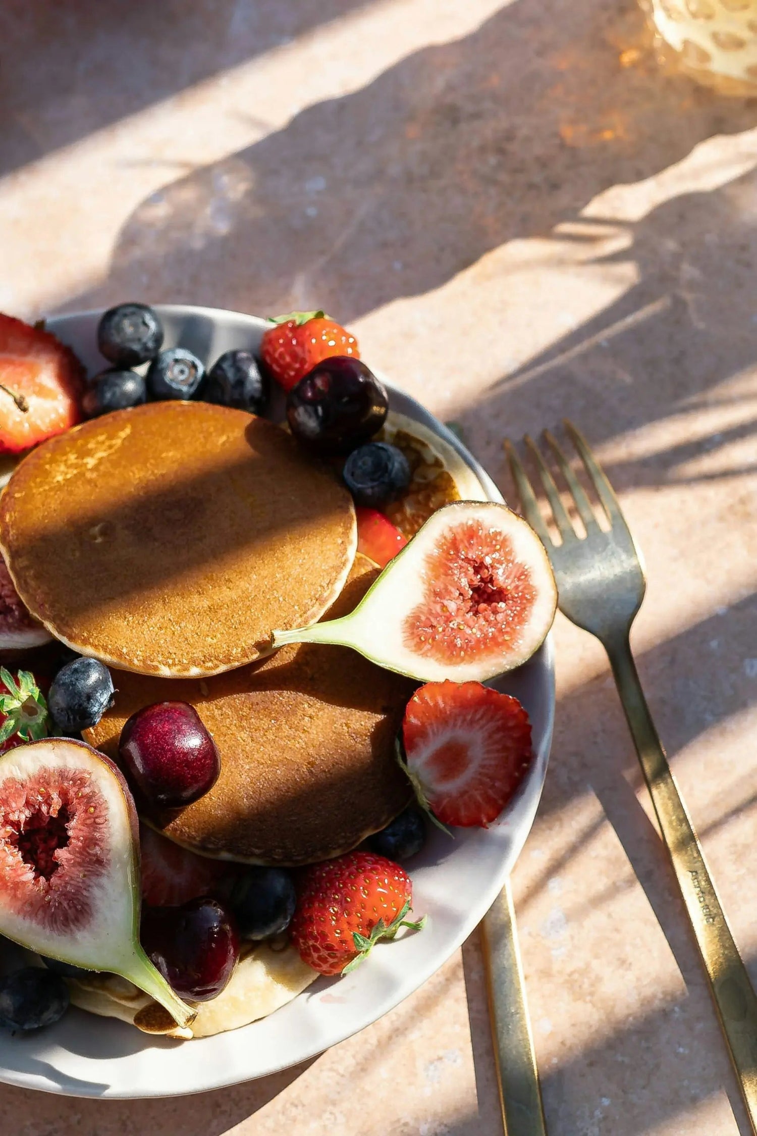 Plate of golden pancakes topped with figs, strawberries, blueberries and cherries beside gold fork and knife on a sunlit table