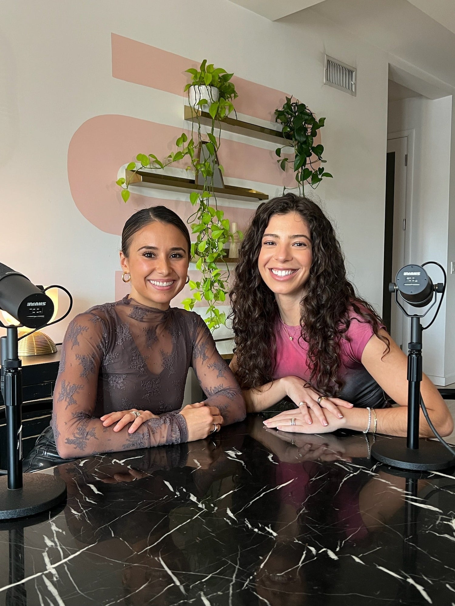 Two women smiling while recording a podcast at a marble table with microphones and hanging potted plants behind them