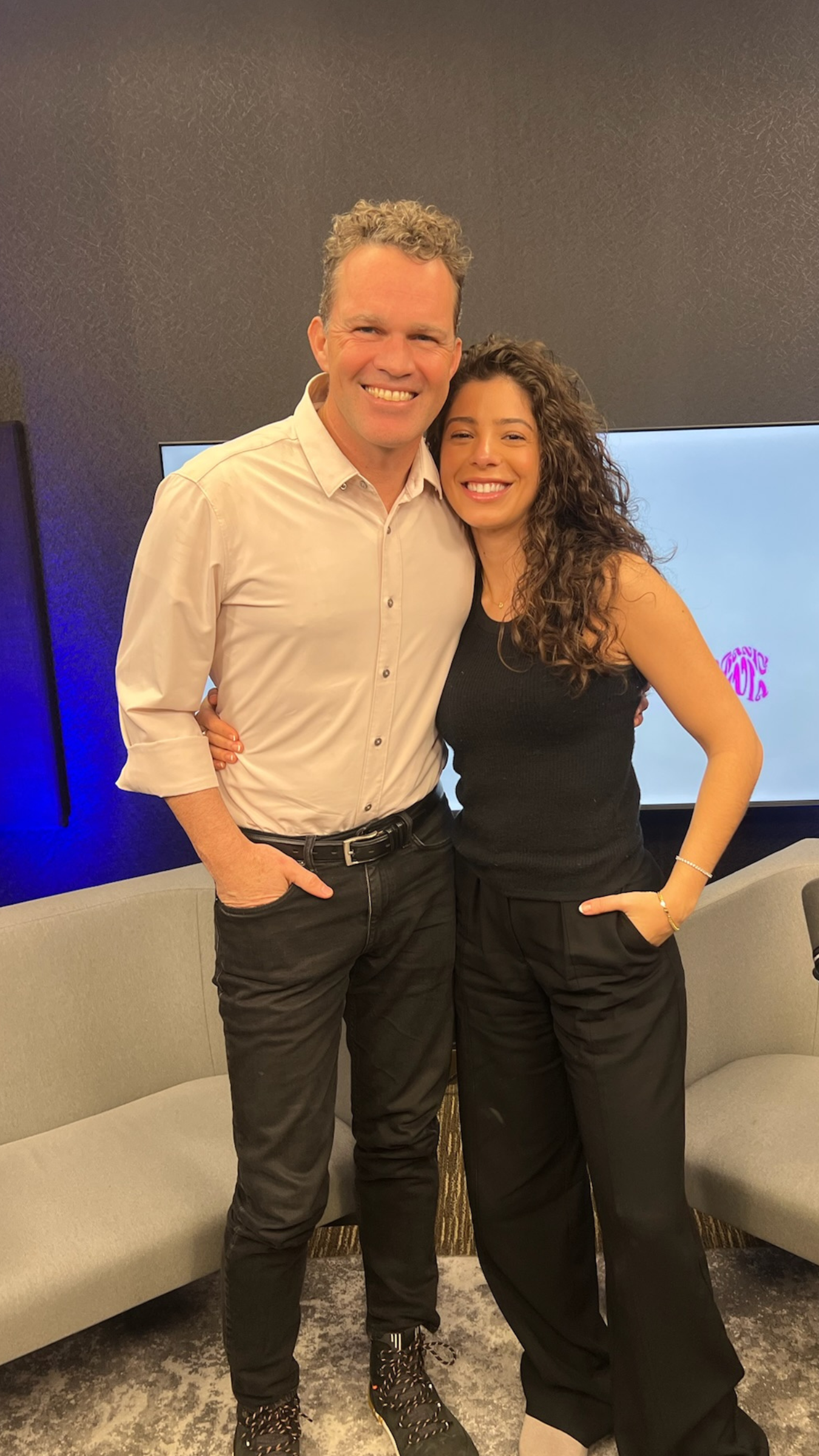 Two people smiling and embracing in a podcast studio with a couch and TV in the background