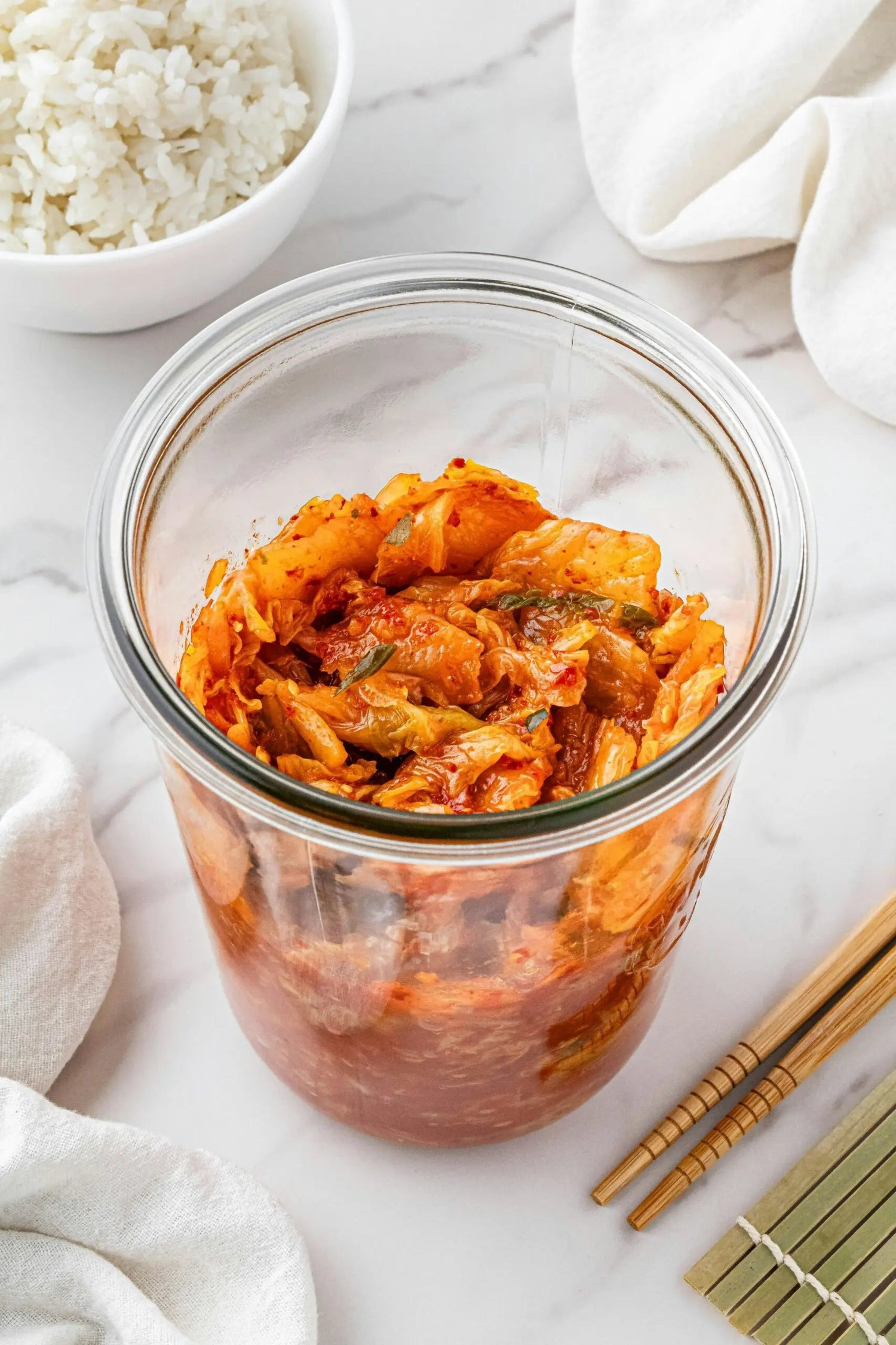 Top-down view of a jar of kimchi showing fermented napa cabbage in spicy red sauce with rice and chopsticks
