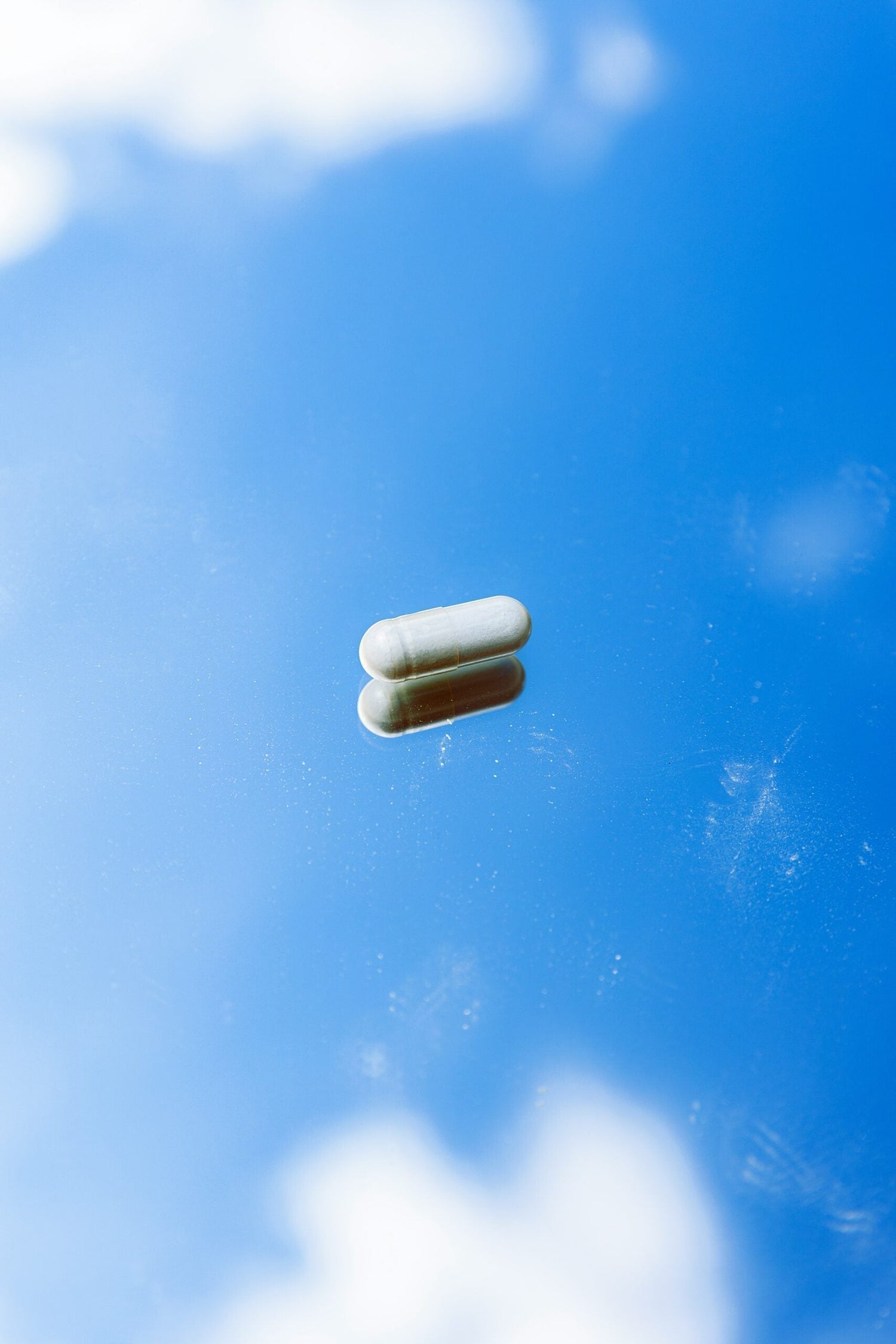 Single capsule resting on a reflective surface with blue sky and cloud reflection