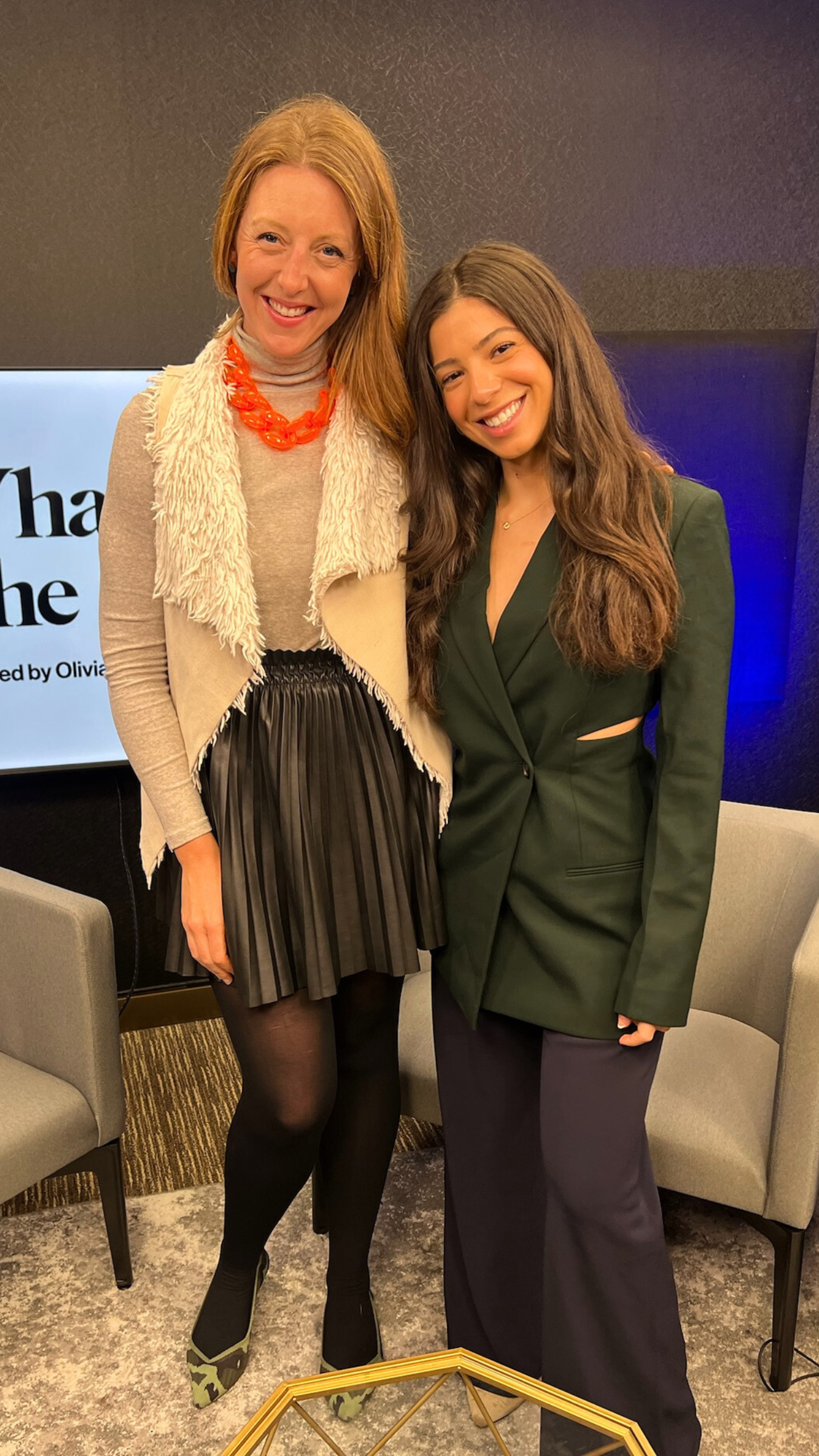 Two women smiling and posing together in a podcast studio with chairs and a screen in the background