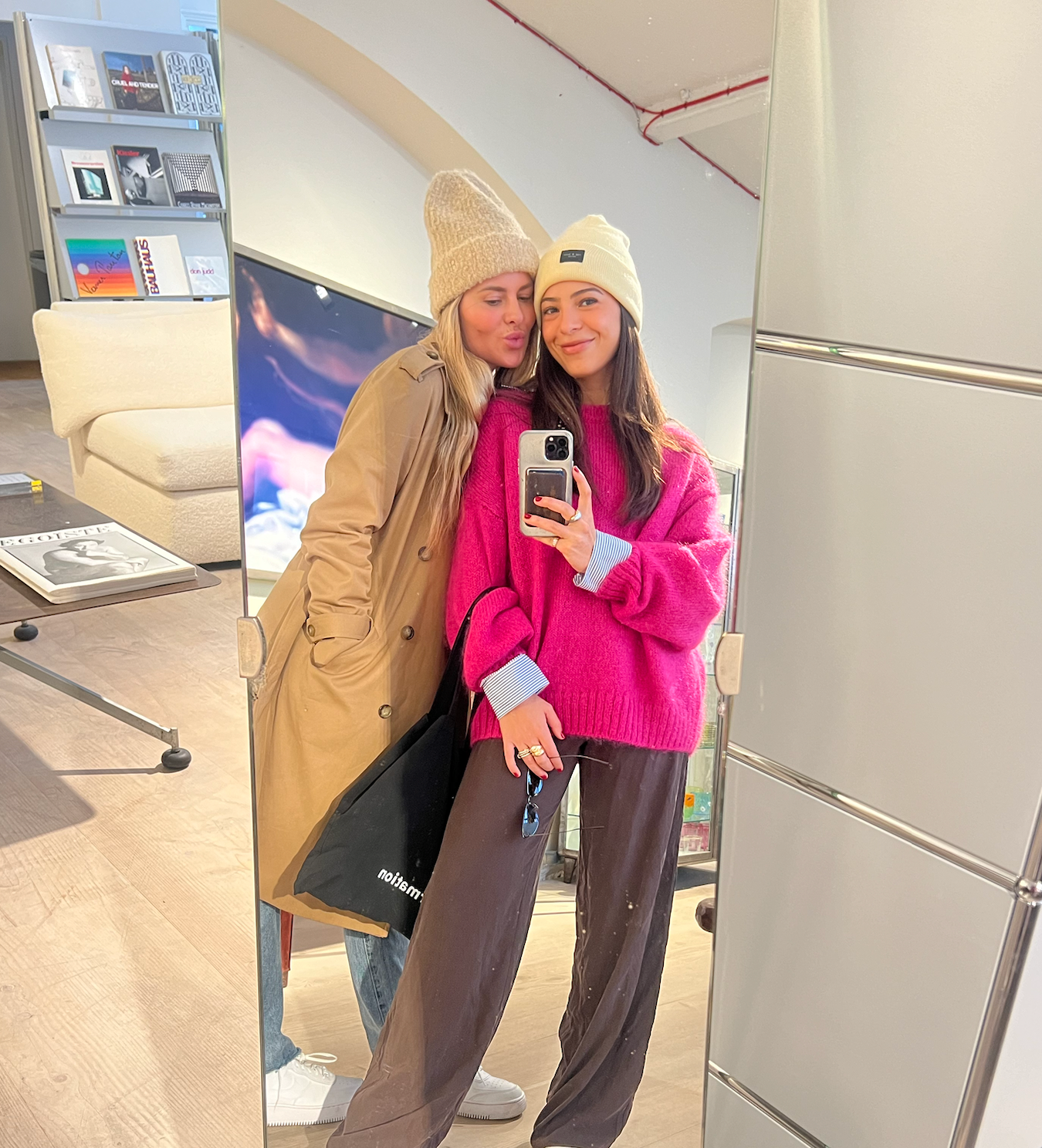 Two women taking a mirror selfie, one in a bright pink sweater holding a phone, the other in a tan coat leaning in