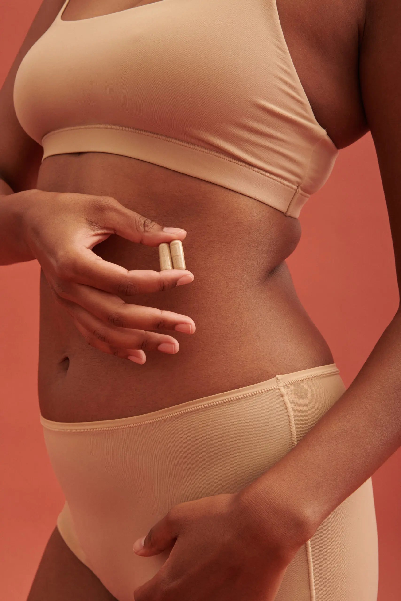 Woman holding two supplement capsules at her lower abdomen, wearing nude undergarments against a warm terracotta background