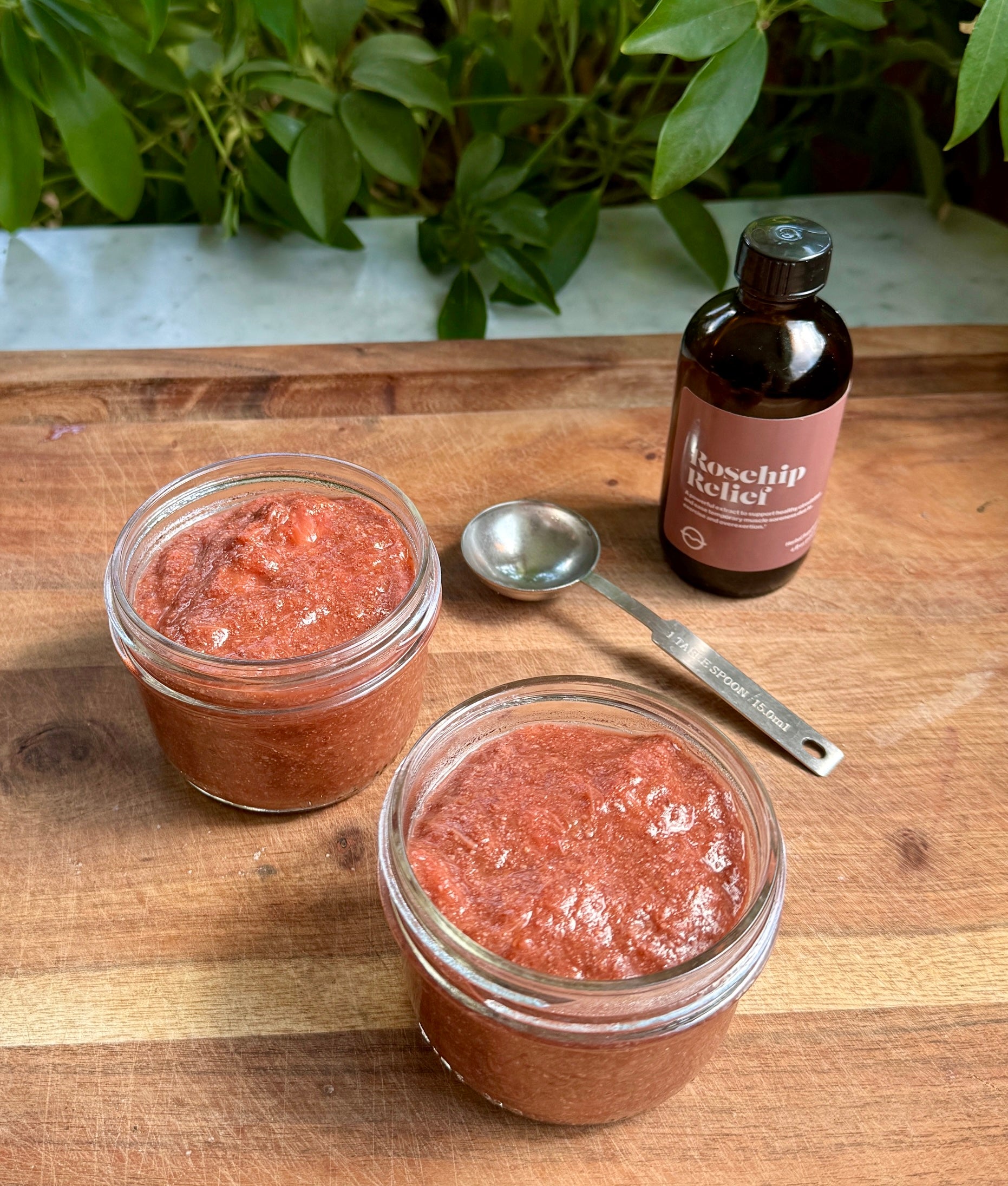 Angled view of two jars of rosehip rhubarb jam with chunky texture, a measuring spoon, and an amber bottle on a wooden board
