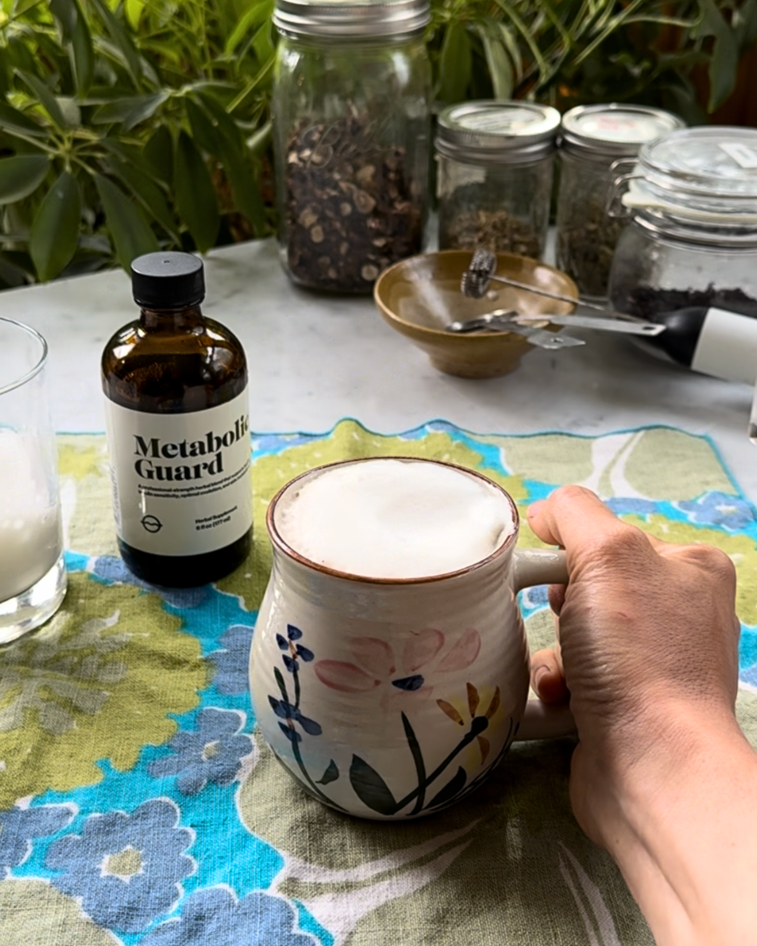 Hand holding a ceramic mug of frothy chai latte on a cloth, bottle labeled Metabolic Guard and jars in background