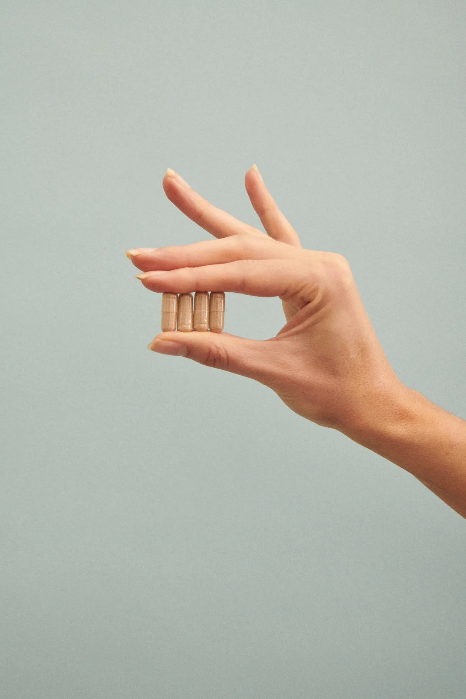 Close-up of four Vitamin B6 capsules held between fingers showing capsule size and beige powder filling against a pale blue-gray background