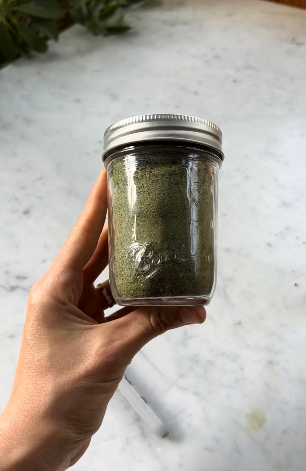 Handheld front view of a mason jar of green herbal salt showing fine ground texture on a white marble countertop