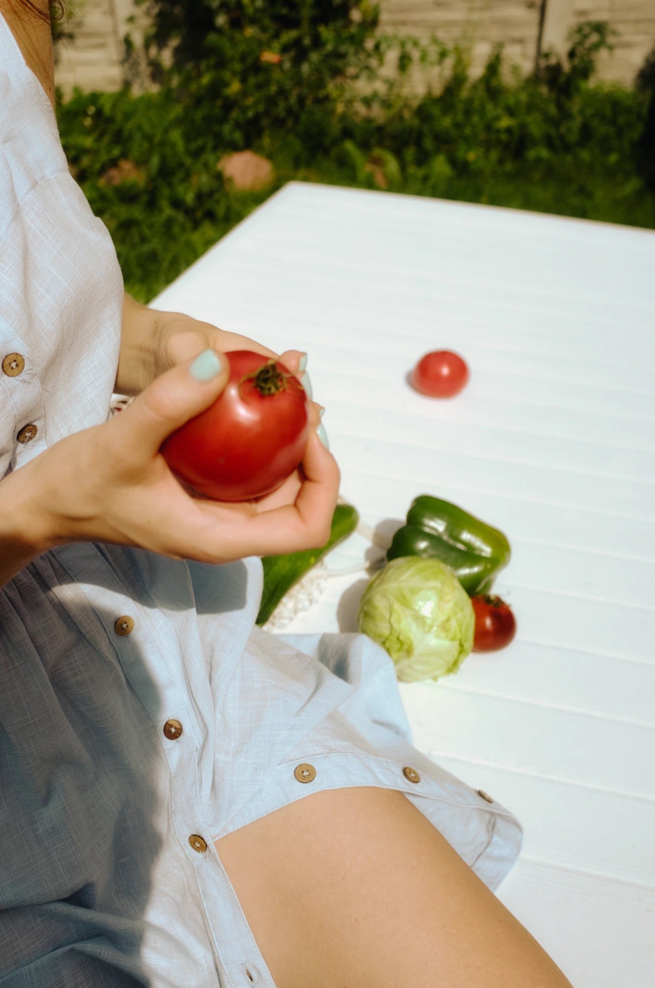 Person holding a ripe tomato at a white table with lettuce and a green pepper in a sunny backyard