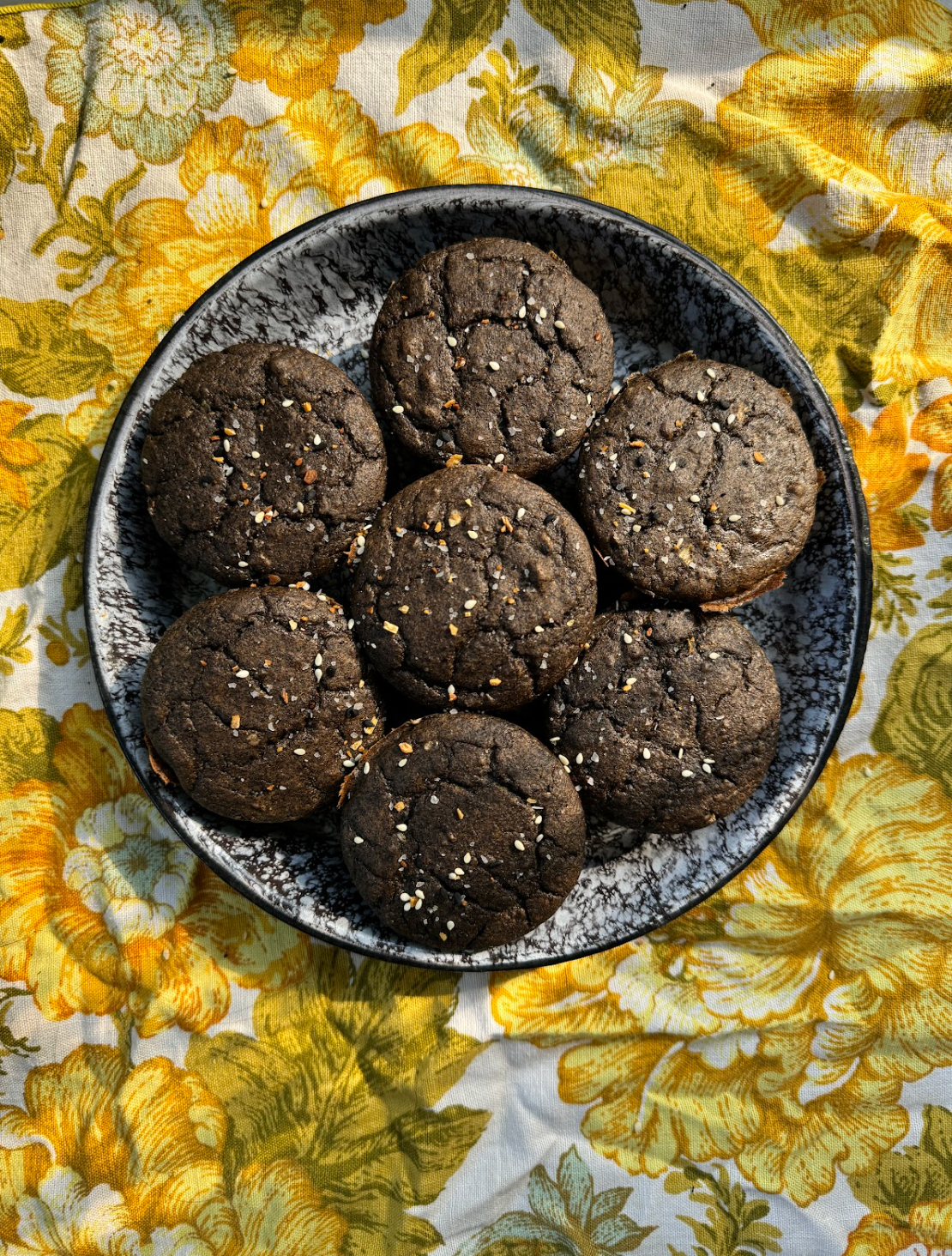 Overhead view of seven savory breakfast muffins in a speckled bowl showing cracked tops and sesame seed seasoning on a yellow floral cloth