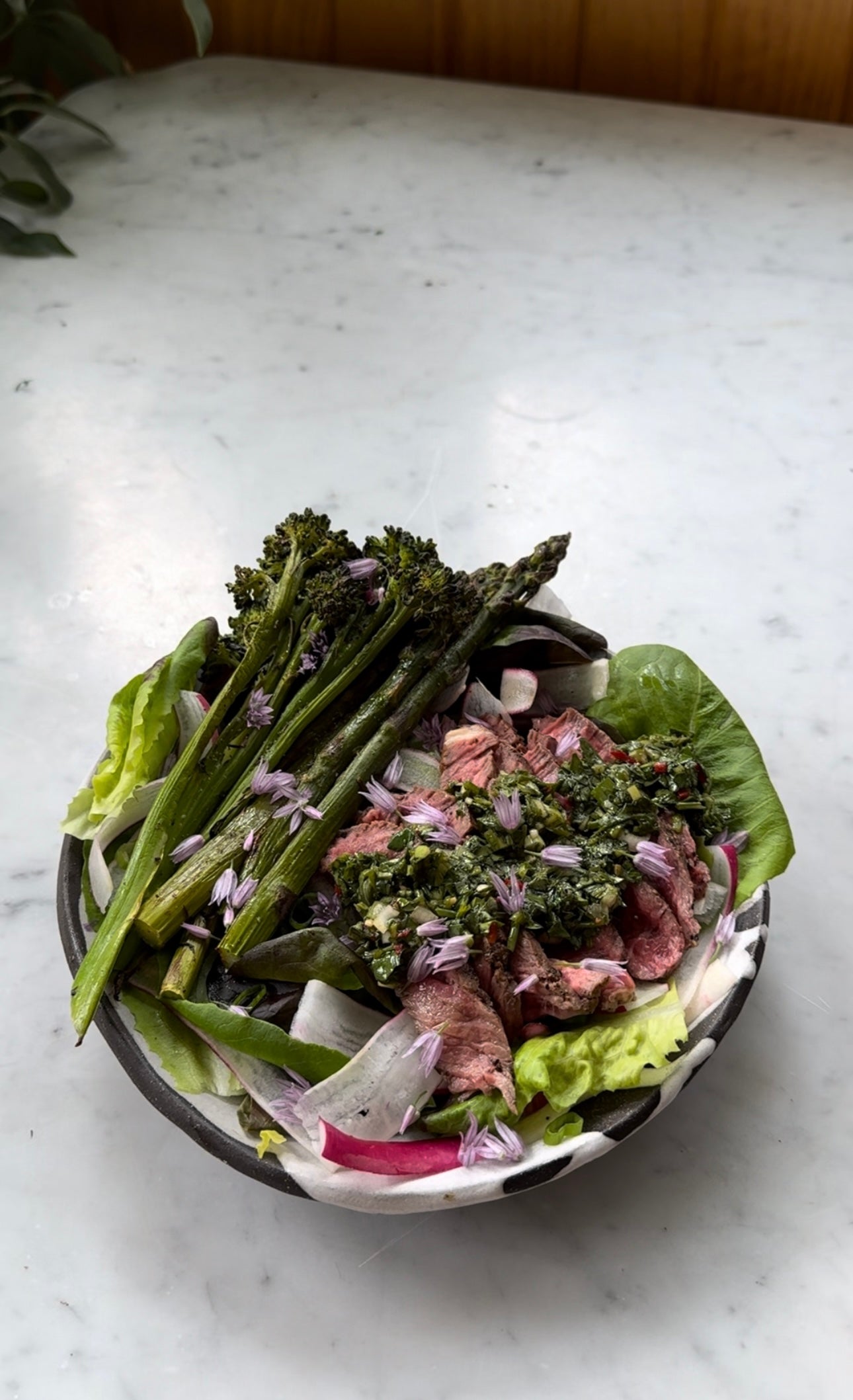 Bowl of sliced steak with herby chimichurri, grilled asparagus, mixed lettuces and radish slices on a marble tabletop