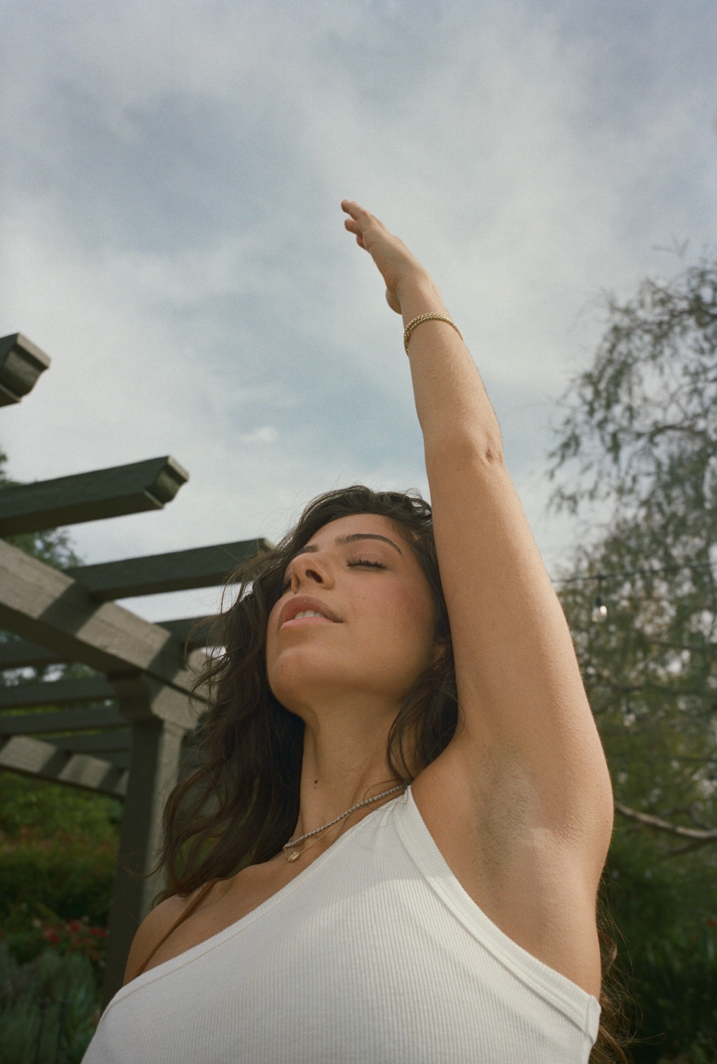 Woman with long dark hair raising one arm toward the sky in a sunlit garden