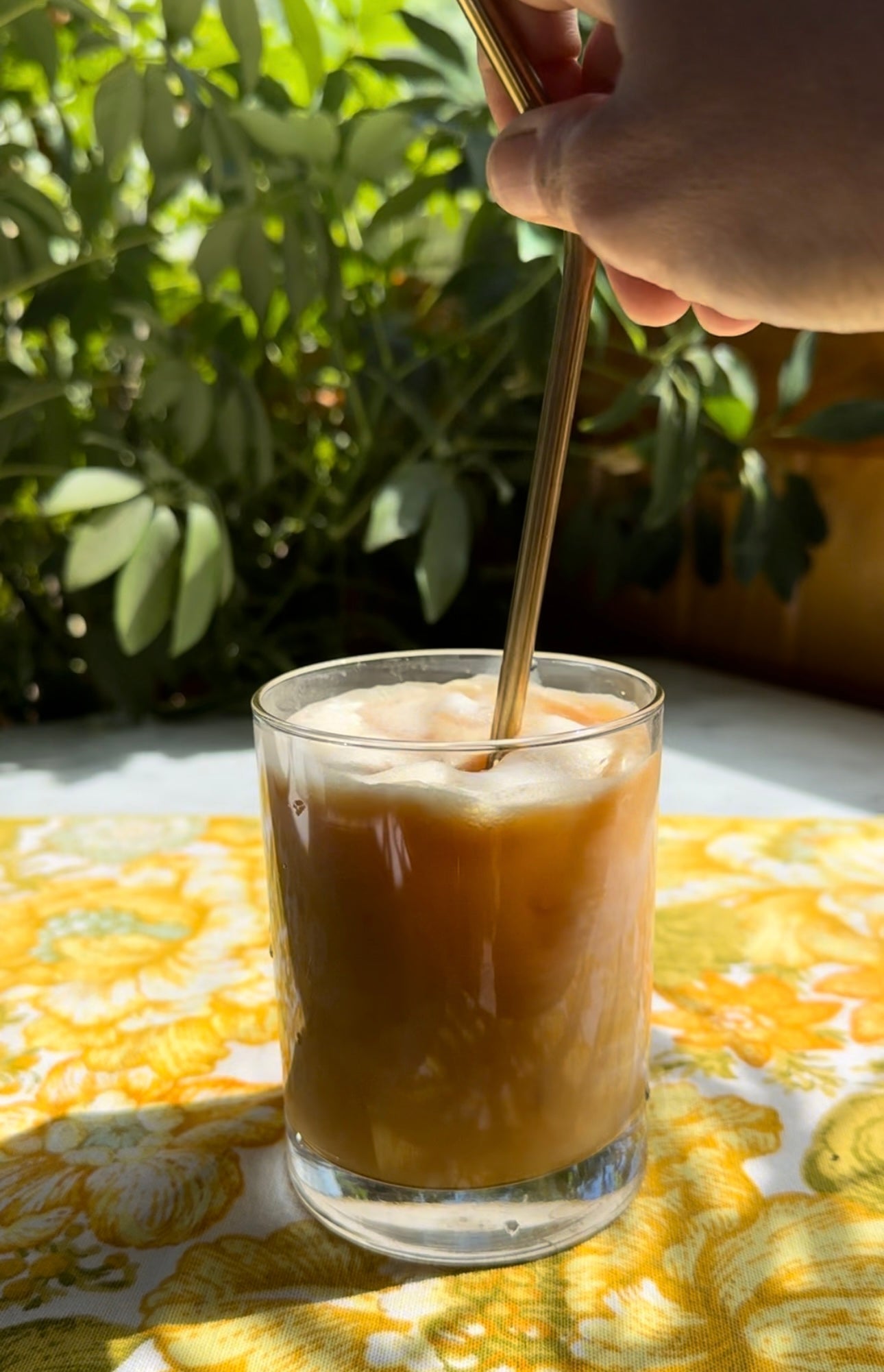 Iced chicory latte being stirred with a brass straw in sunlight on a yellow floral tablecloth, leafy plant in the background