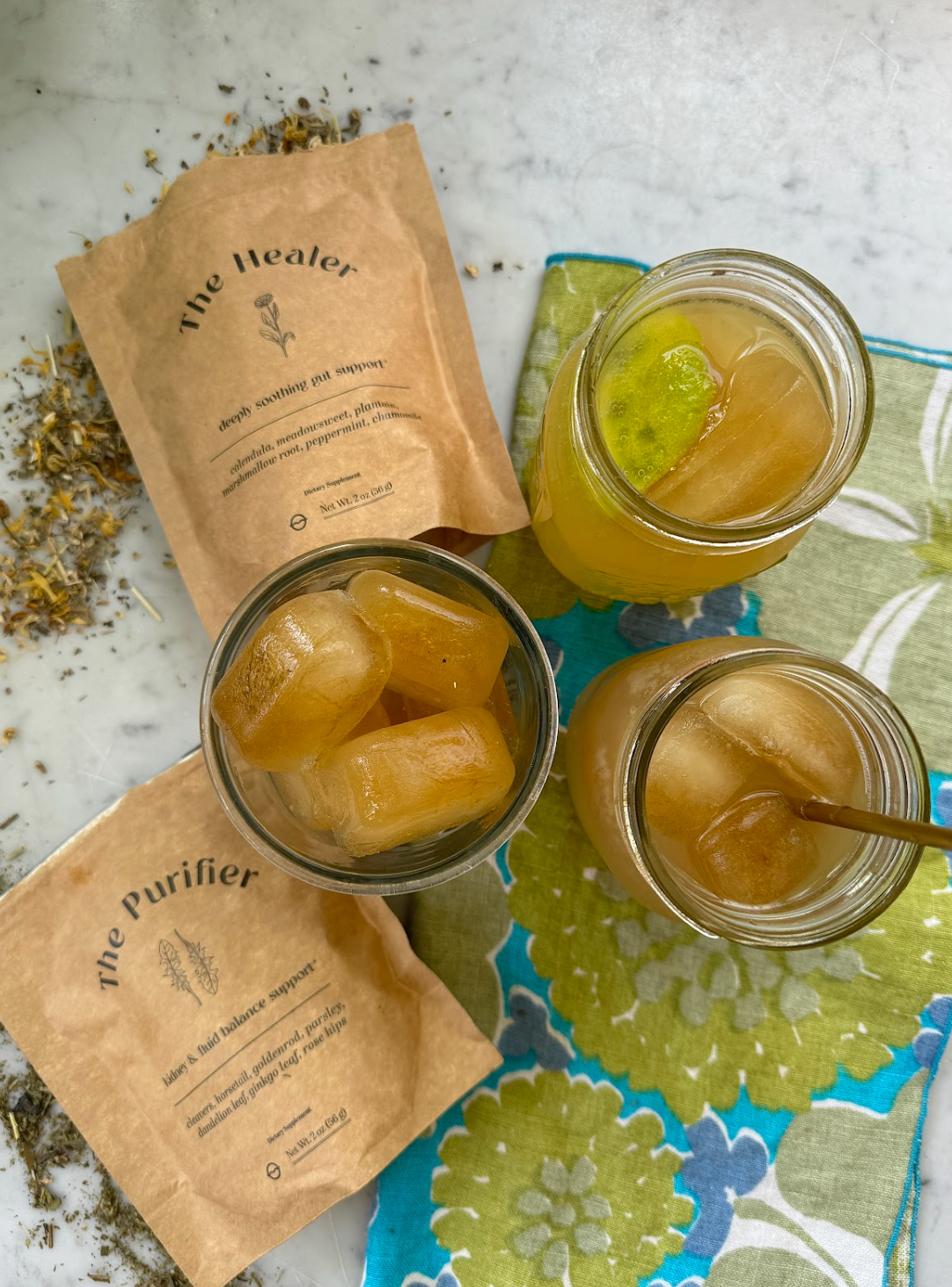 Top-down view of herb-infused ice cubes chilling iced tea in mason jars beside kraft packets labeled The Healer and The Purifier