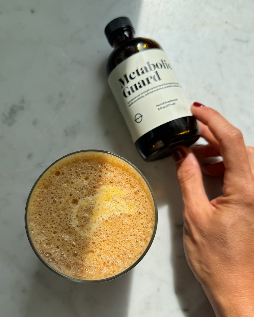 Hand holding a Metabolic Guard bottle beside a glass of frothy pumpkin cold foam on a marble countertop lit by sunlight