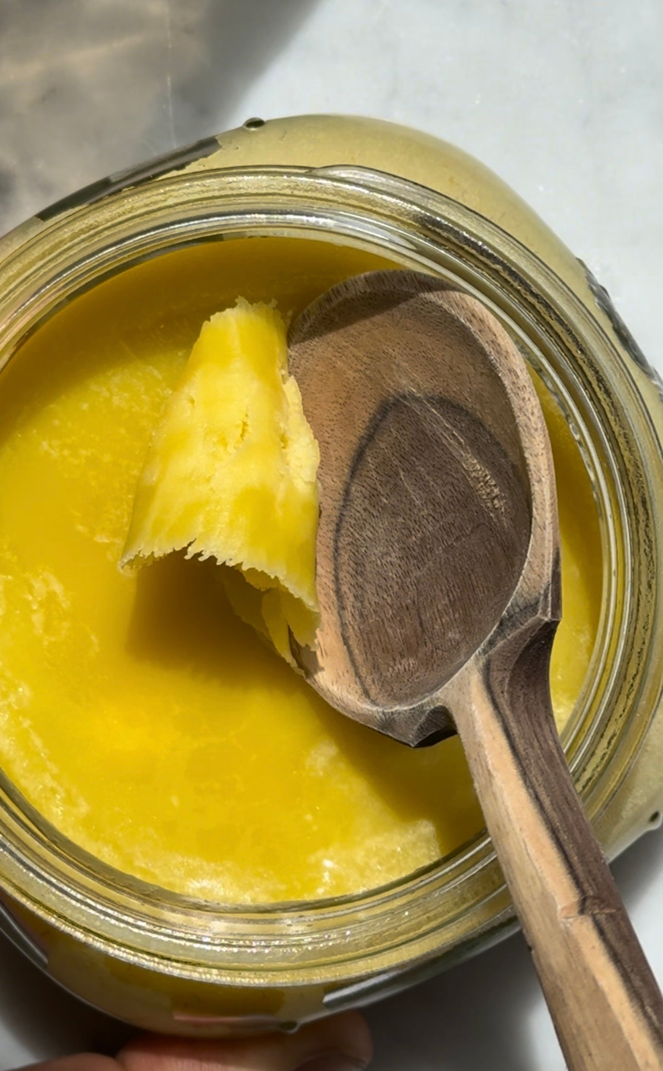Top-down view of jar of herb infused ghee showing creamy yellow texture with wooden spoon on marble surface