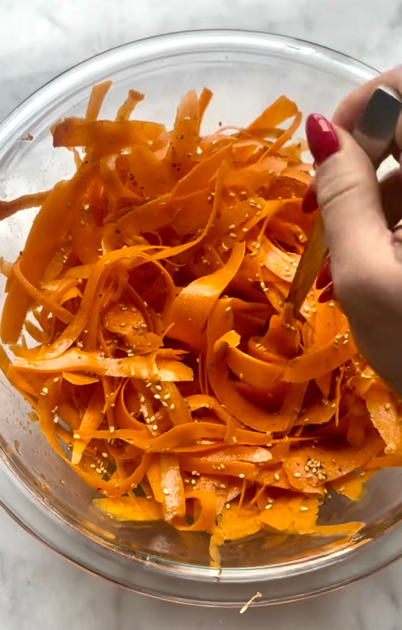 Shaved raw carrot salad being tossed with a fork in a glass bowl sprinkled with sesame seeds on a marble countertop