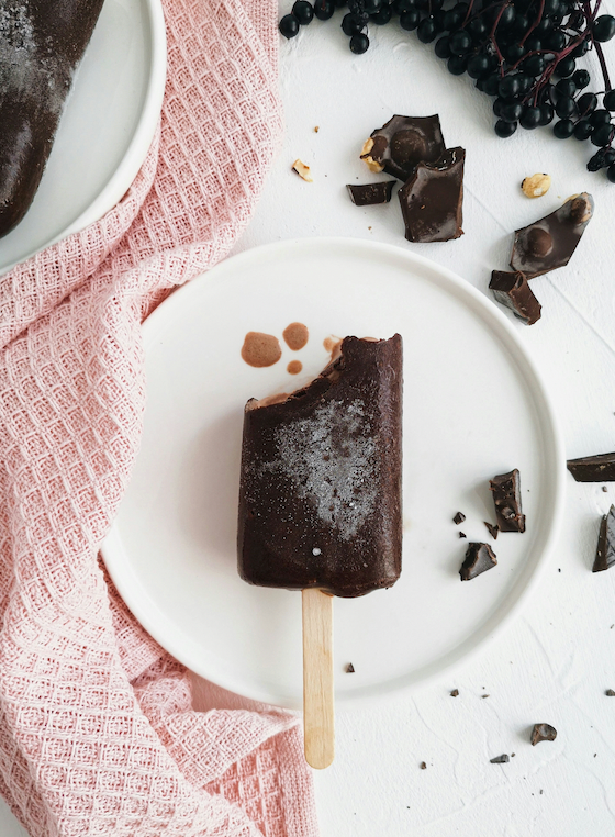 Chocolate-coated popsicle with a bite taken, on a white plate with broken chocolate pieces and a pink textured towel