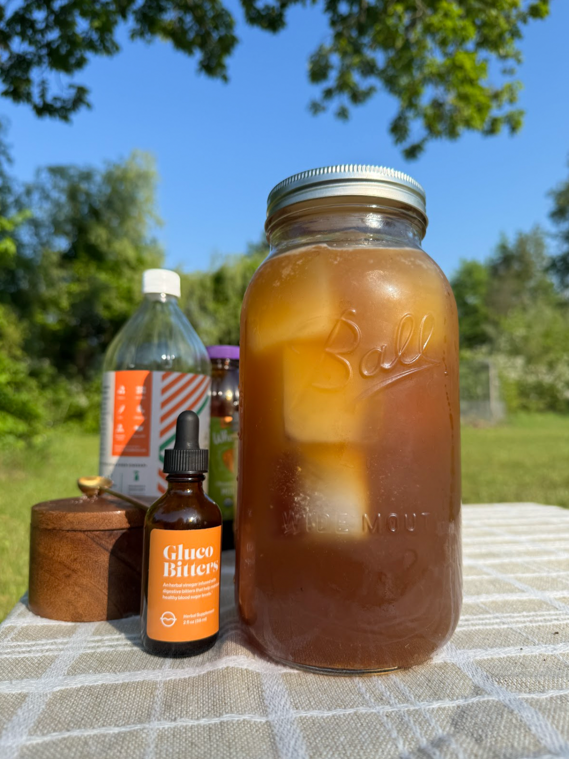 Mason jar of iced switchel beside a bottle of Gluco Bitters and other ingredients on a picnic table outdoors
