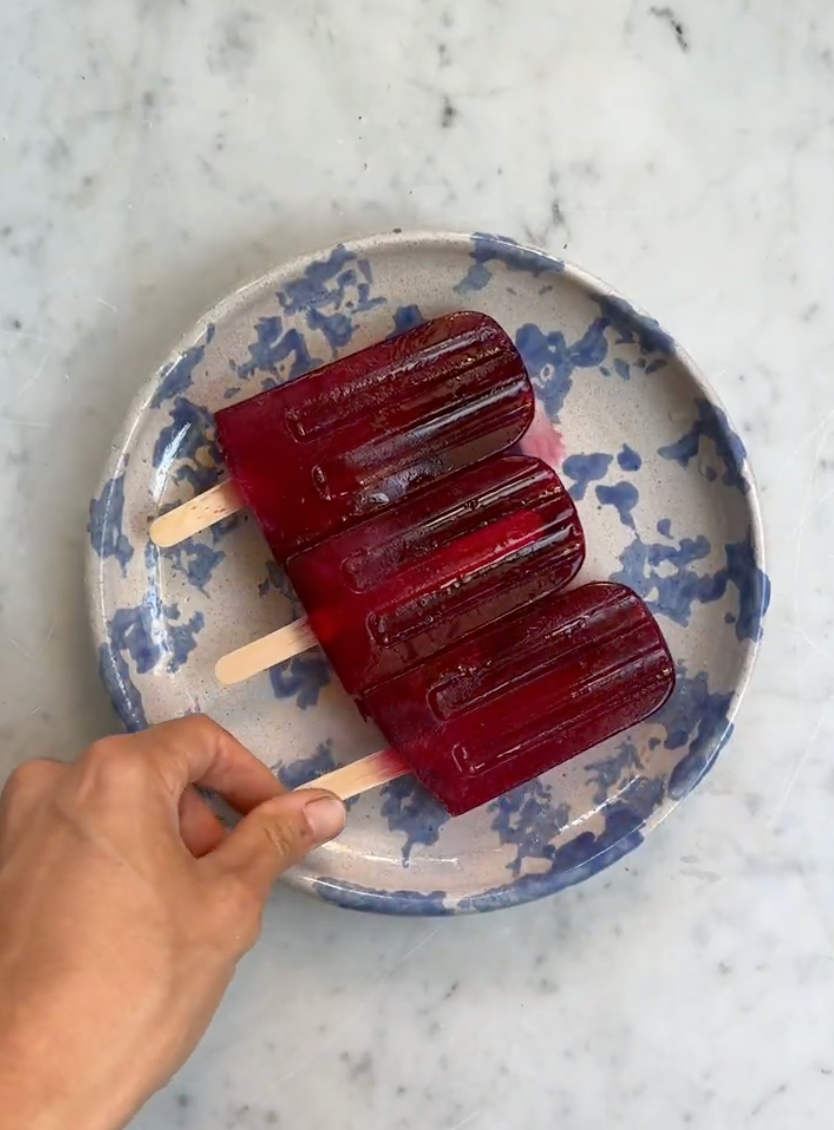 Three deep-red herbal popsicles on a blue-speckled ceramic plate, a hand picking one up on a marble countertop