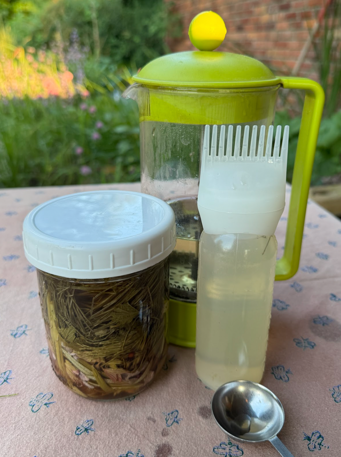 Glass jar of steeping herbs with applicator bottle and green French press on a pink floral tablecloth in a garden