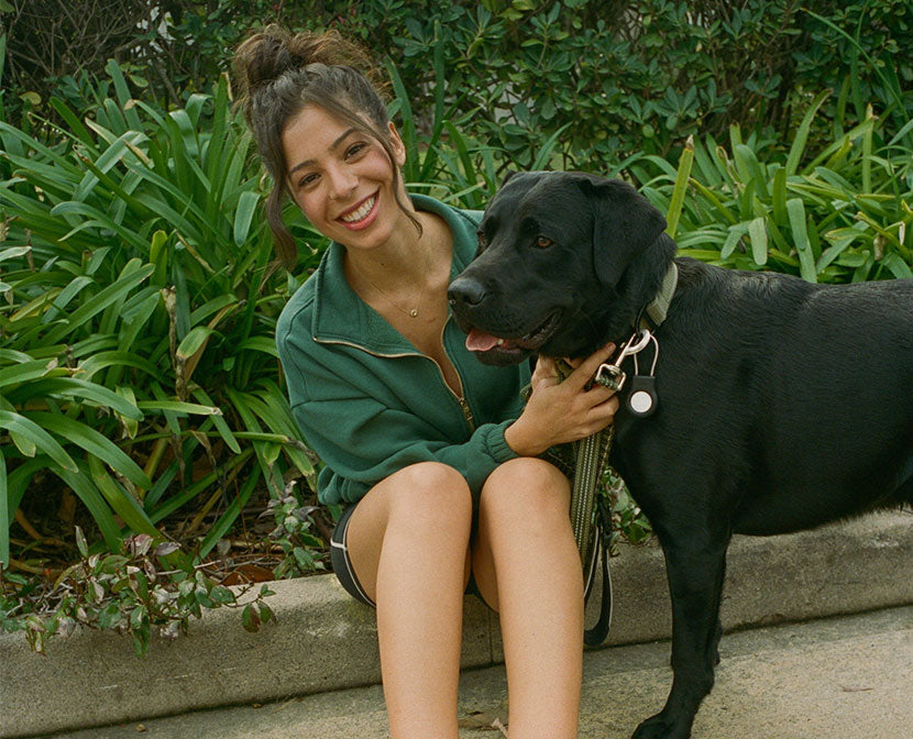 Woman sitting on a curb smiling while holding a black Labrador on a leash with green plants behind them