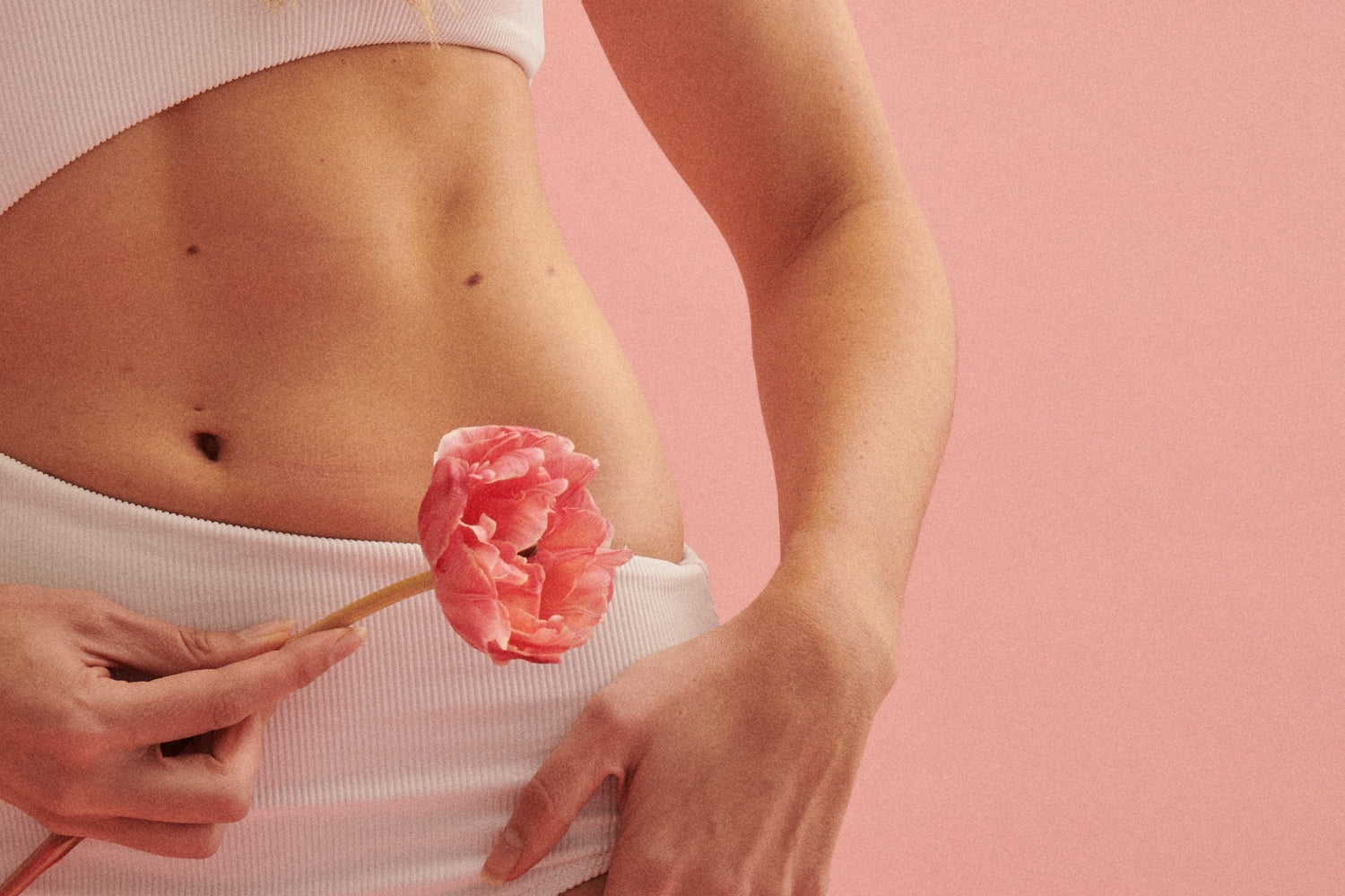 Close-up of a woman’s midsection holding a pink tulip against her lower abdomen with a solid pink background