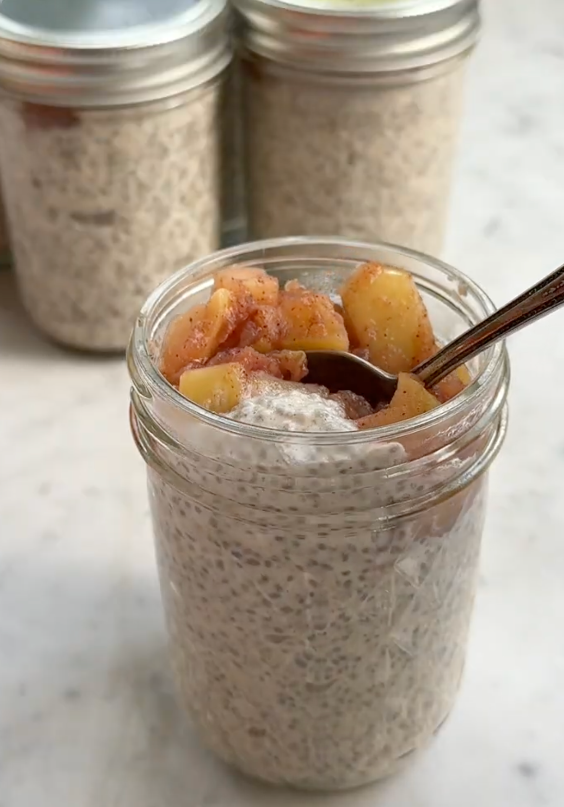 Caramel apple chia pudding in a glass jar topped with cinnamon-spiced apple pieces and a spoon, chia seed jars blurred in the background