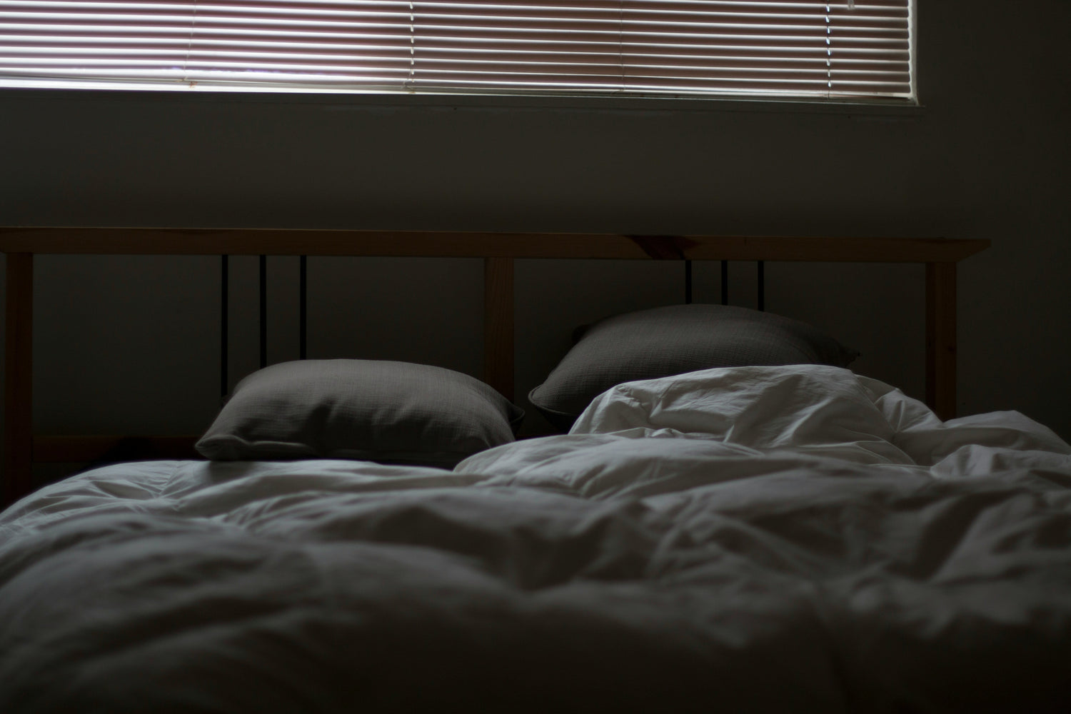 Unmade bed with white duvet and two gray pillows beneath a window with closed blinds in a dim bedroom