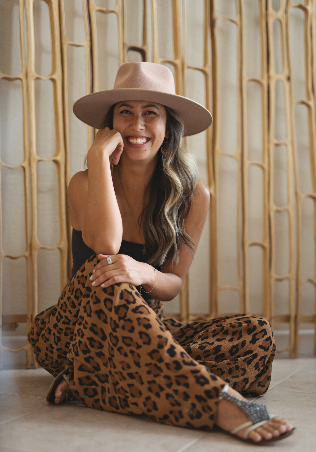 Smiling woman sitting cross‑legged on a tile floor wearing a wide‑brim hat and leopard‑print pants in front of a wooden room divider