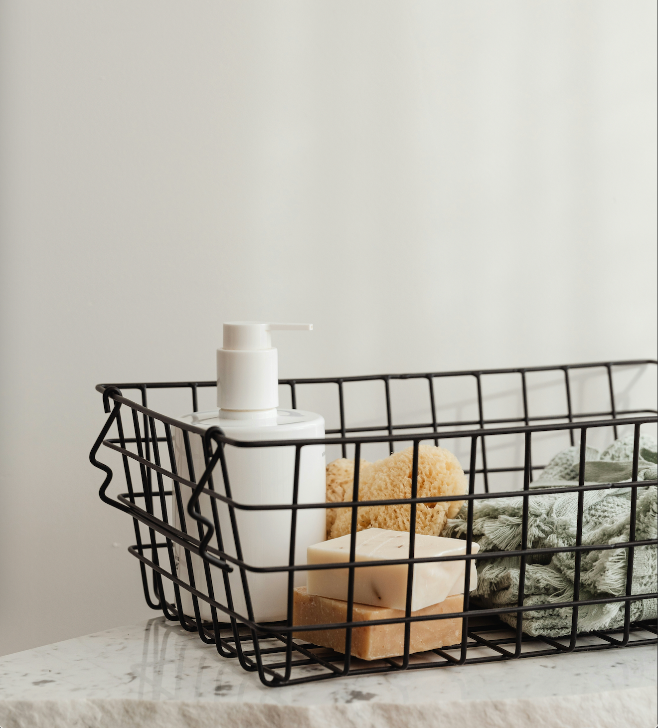 Wire basket holding a pump bottle, two stacked soap bars, a natural sponge, and a folded green towel on a marble countertop