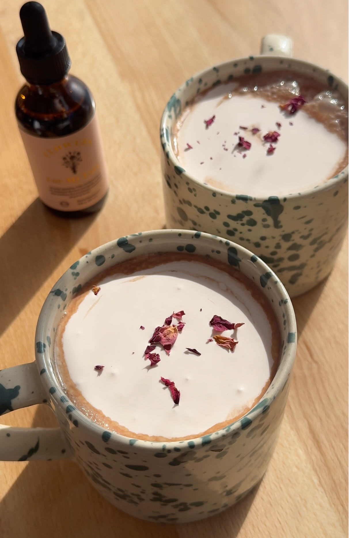 Two mugs of rose hot cocoa topped with frothed milk and dried rose petals on a sunlit wooden table next to a dropper bottle