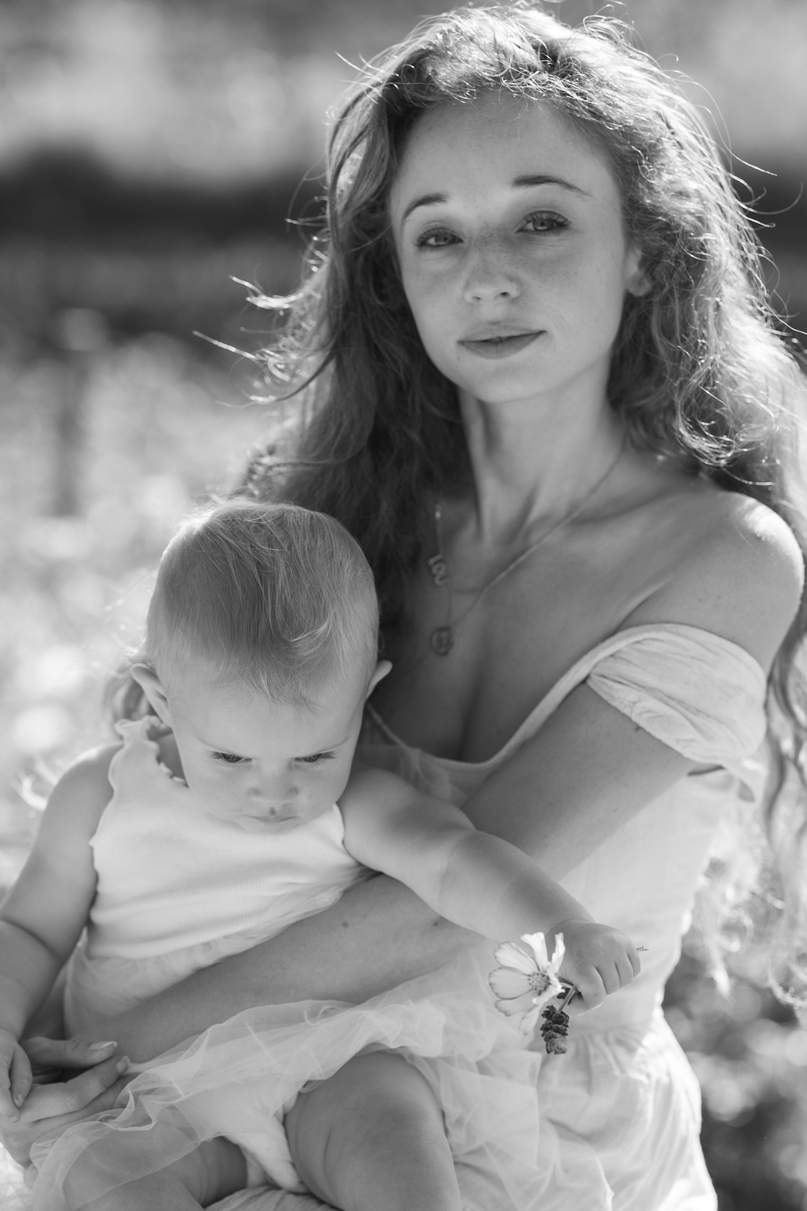 Mother holding an infant in her lap outdoors, mother looking at camera while baby reaches for a flower
