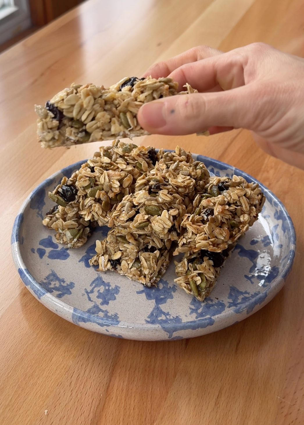 Hand picking up a homemade granola bar from a blue speckled plate on a wooden table, showing oats, pumpkin seeds and dried cherries