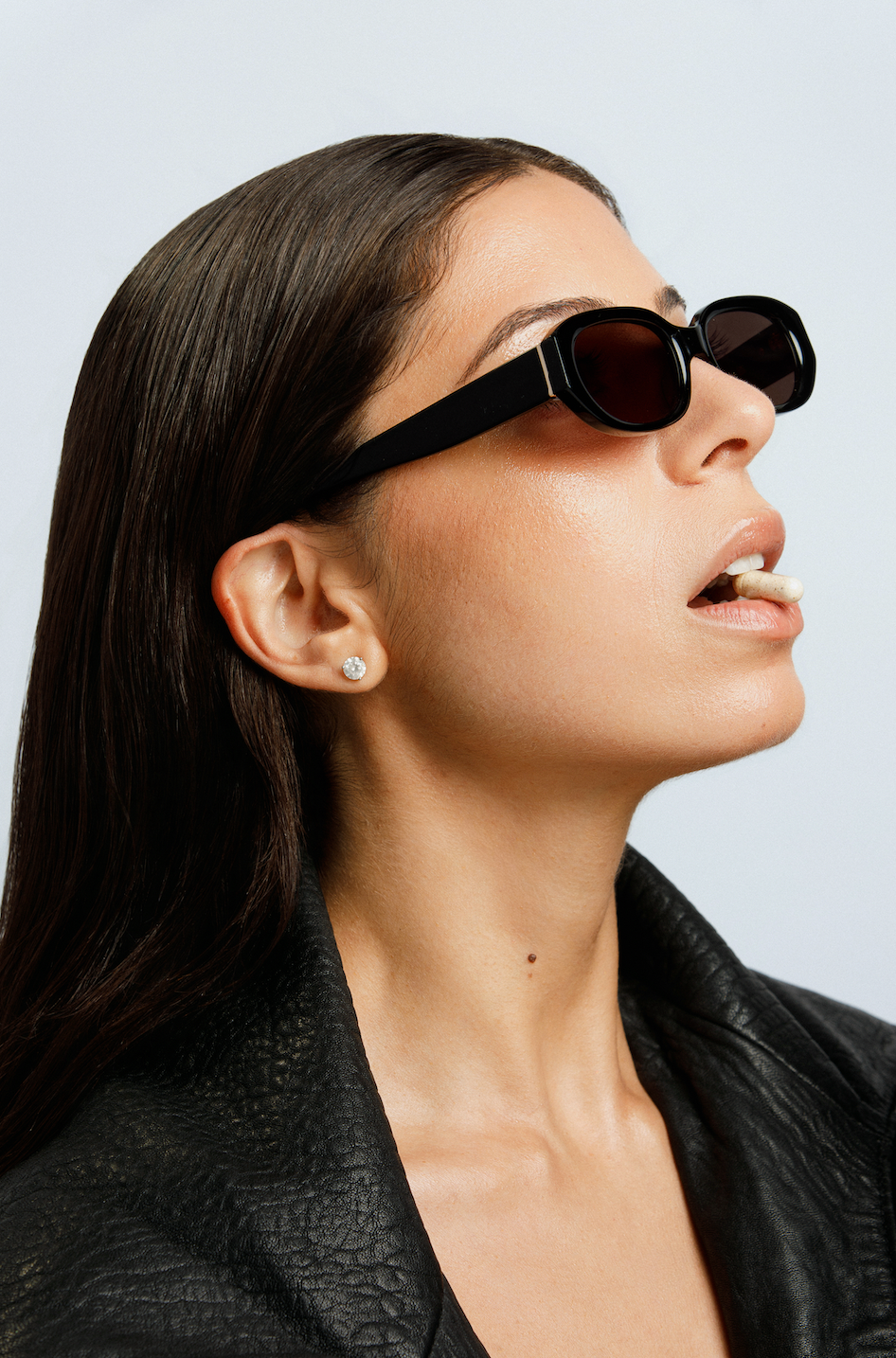 Woman with long dark hair wearing black sunglasses and a leather jacket holding a supplement capsule between her teeth, head tilted back against a pale background