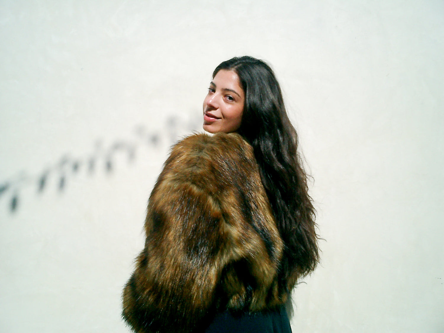 Woman with long dark hair looking over her shoulder wearing a brown faux-fur jacket against a pale wall