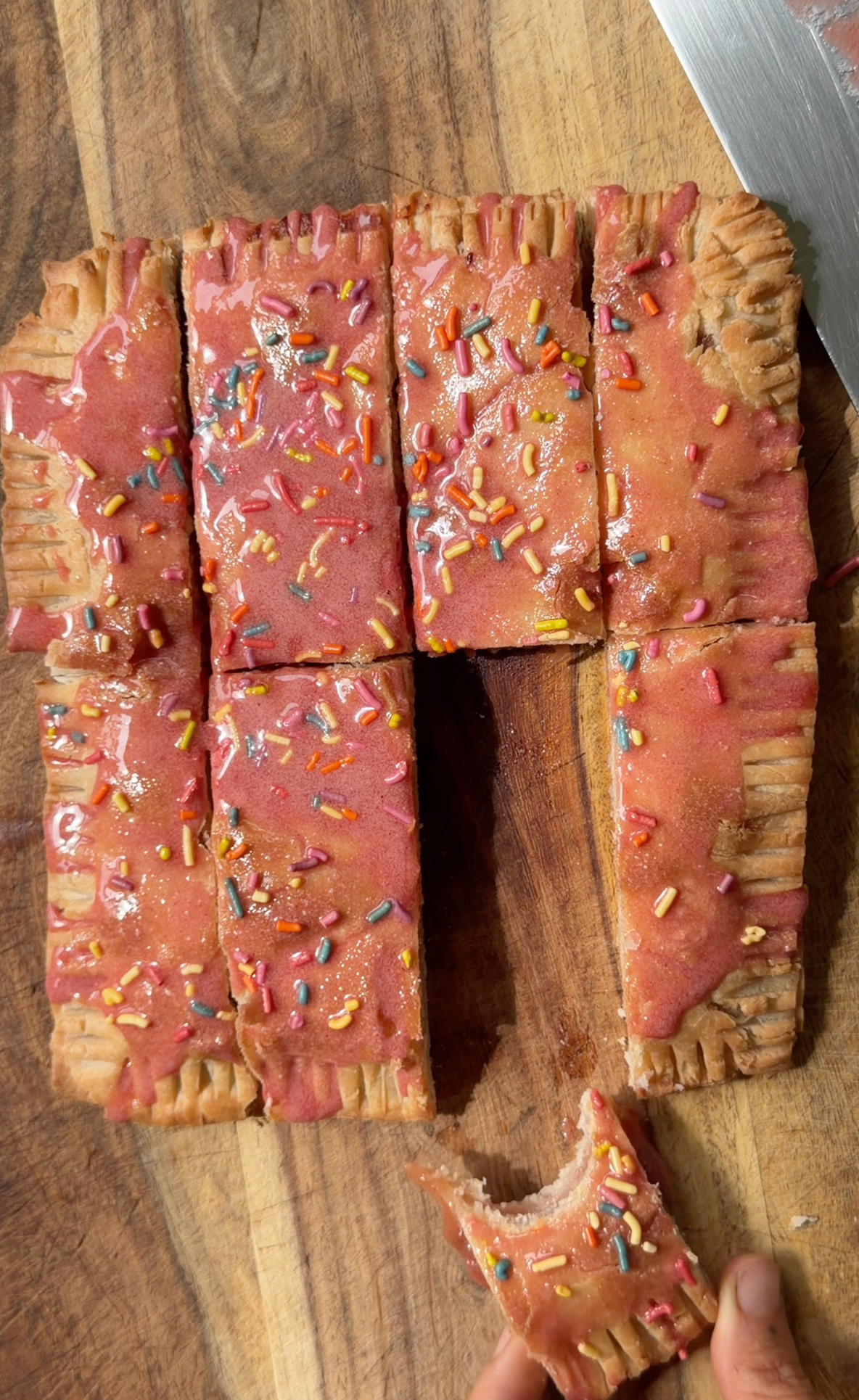 Strawberry rhubarb pop‑tart being held after a bite, glazed with pink icing and rainbow sprinkles on a wooden cutting board