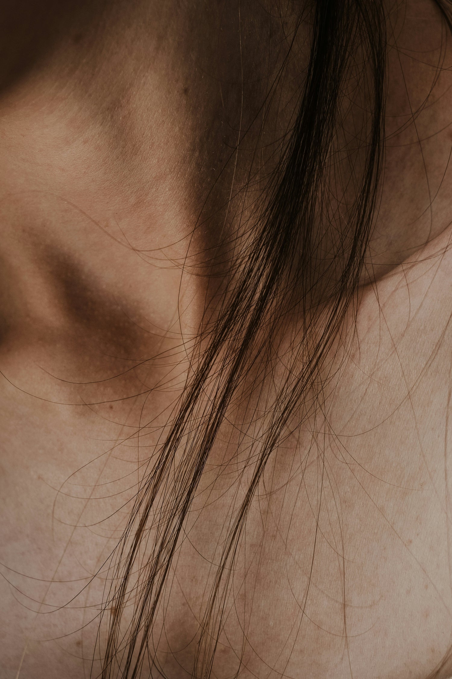 Close-up of a person’s neck and collarbone with long dark hair strands in warm sepia lighting