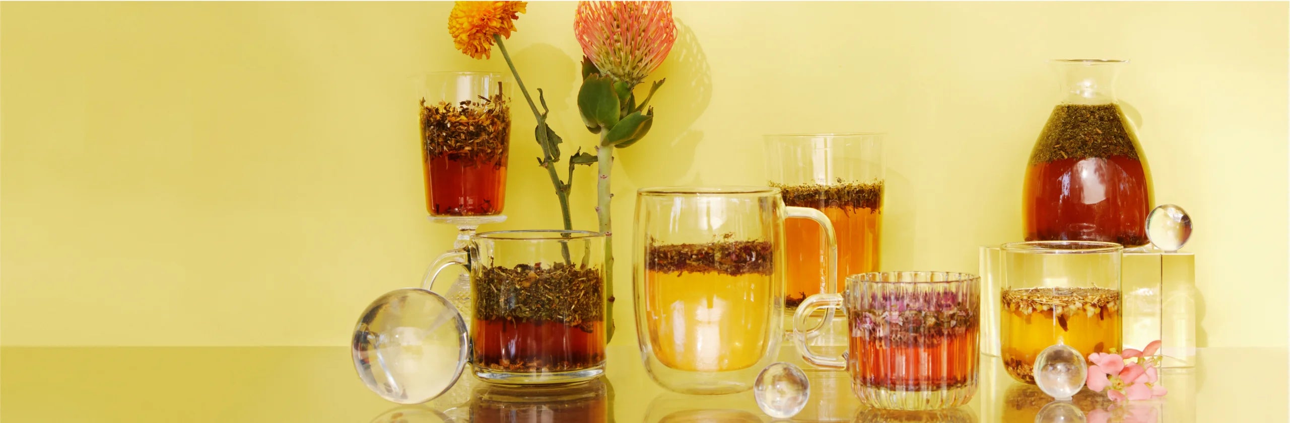 Glass cups and a carafe of steeping herbal teas arranged on a reflective surface against a warm yellow background